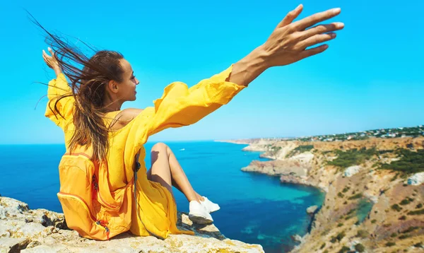 Primer plano retrato de mujer viajera bronceada sentada en la playa de roca superior con las manos levantadas, vestido amarillo brillante y cabello soplando en el viento . — Foto de Stock