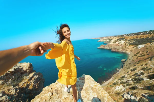 Porträt einer glücklichen, unbeschwerten Frau, die die Hand ihres Freundes hält, auf einer Klippe steht, ein leuchtend gelbes Kleid trägt und die Haare im Wind wehen. folge mir. — Stockfoto