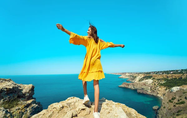 Rear view of taned traveler woman standing on top rock beach with open arms, bright yellow dress and hair blowing in the wind. — kuvapankkivalokuva