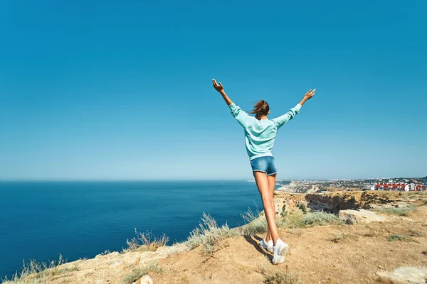 Vista de fondo de la joven sin preocupaciones de pie con las manos levantadas en el borde del acantilado con hermosas vistas al mar y disfrutar de la naturaleza maravillosa. Retrato completo de la longitud — Foto de Stock