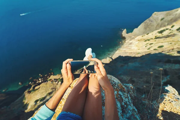 Uçurumun kenarında oturan güzel deniz manzaralı ve akıllı telefon kamerasıyla suyun üstünde bacaklarını çeken ilk kişi.. — Stok fotoğraf