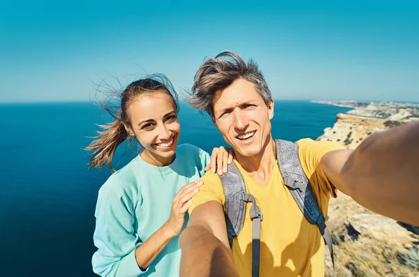 Young man and woman tourist making selfie and smile to the camera on cliff edge with beautiful sea view and enjoying wonderful nature. — kuvapankkivalokuva