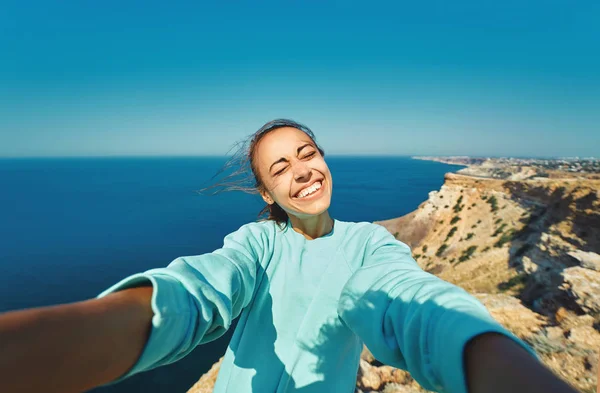 Nuori naispuolinen matkustaja tekee selfien kalliolla reunalla kauniilla merinäköalalla, nauraen ja hymyillen kameralle, tehden hauskat kasvot . — kuvapankkivalokuva