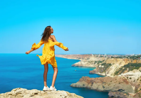 Retrato completo de la mujer viajero bronceada que se encuentra en la playa de la roca superior con los brazos abiertos, vestido amarillo brillante y el cabello soplando en el viento.. — Foto de Stock