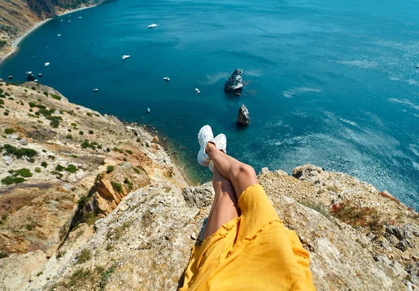 Uçurumun kenarında oturan sarı elbiseli bronzlaşmış kadın bacakları ve deniz manzarası inanılmaz.. — Stok fotoğraf