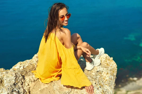 Bright portrait of happy woman in bright yellow dress and red sunglasses sitting on cliff edge against amazing seascape. — kuvapankkivalokuva