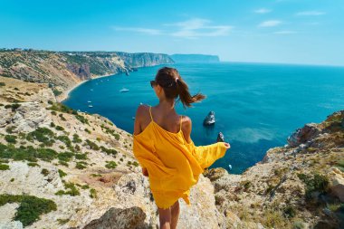 Bronzlaşmış gezgin kadının uçurumun kenarında dikilip, rüzgarda esen parlak sarı elbisesiyle poz verişi..