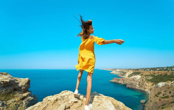 Mujer viajero bronceada que se encuentra en la cima de la playa de roca y da la vuelta, vestido amarillo brillante y el cabello soplando en el viento.. — Foto de Stock