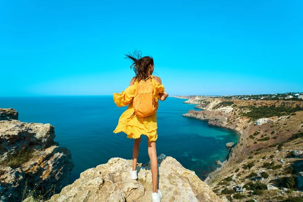 Vista panorámica de la mujer viajero de pie en la playa de roca superior con mochila, vestido amarillo brillante y el cabello soplando en el viento.. — Foto de Stock