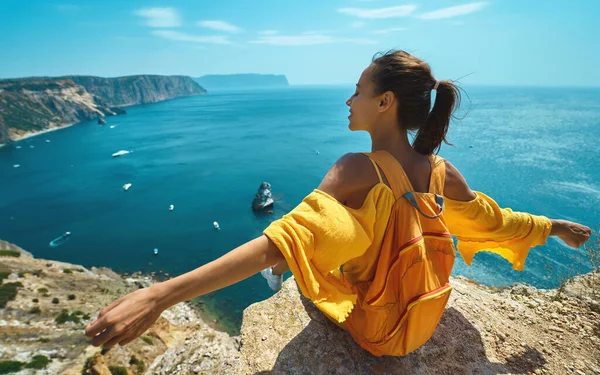 Vista trasera mujer viajero en vestido amarillo brillante con mochila sentado en el borde del acantilado con los brazos abiertos contra increíble paisaje marino . — Foto de Stock
