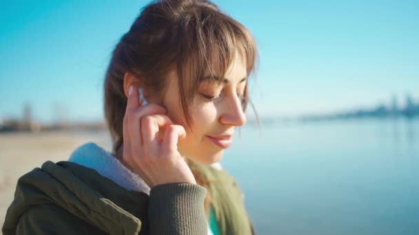 Close-up portret jonge mooie vrouw met behulp van draadloze oortelefoons buiten. — Stockvideo