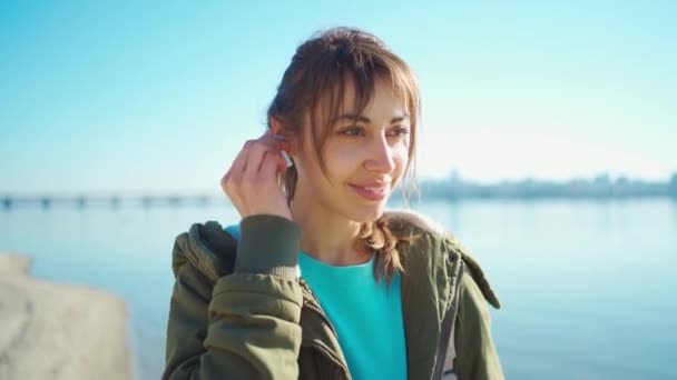 Attraktive Frau steckt drahtlosen Kopfhörer ins Ohr, macht doppelten Wasserhahn und lächelt ruhig und selbstbewusst. — Stockvideo