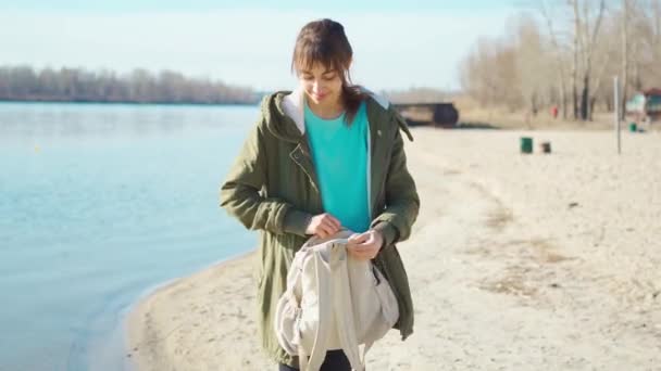 Jovencita mujer alegre poniendo una mochila pequeña y caminando al aire libre a lo largo de la orilla del mar en la playa de arena.. — Vídeos de Stock