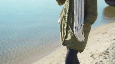 Yeşil ceketli ve beyaz atkılı kadın, güneşli bahar gününde kumlu sahil şeridinde yürüyor.