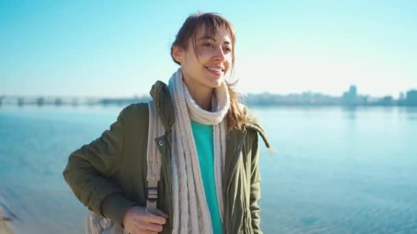 Attraktive lächelnde junge brünette Frau, die einen kleinen Leinenrucksack anzieht und draußen am Strand spazieren geht. — Stockvideo