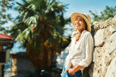 Hasır şapkalı mutlu kadın turist, Tayland 'da tatilde eski taş duvarın yanında duran beyaz gömlek, gülümseyerek ve tek başına seyahatin tadını çıkarıyor..