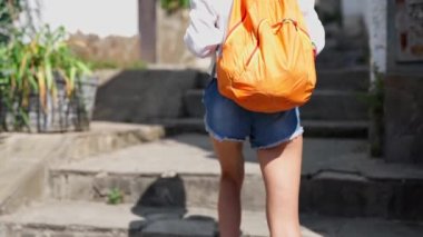 Arka plan kamerası, bronzlaşmış uzun bacaklı, kot şortlu, seksi kadın turisti takip ediyor. Eski dar sokakta merdivenlerden yukarı çıkıyor.