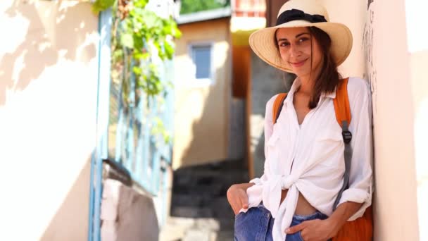 Молодая элегантная туристка в соломенной шляпе, джинсовых шортах, белой рубашке и оранжевом рюкзаке наслаждается прогулкой по узкой улице — стоковое видео