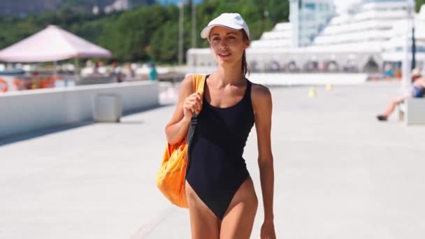 Mooie stijlvolle sexy sportieve vrouw in zwart badpak en witte muts wandelen langs baai aan zee — Stockvideo