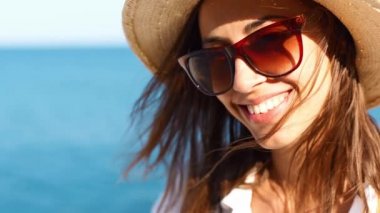 Yakın çekim gülümseyen kadın gezgin hasır şapkalı ve güneş gözlüklü güneşli bir günde deniz kenarında yürüyor.