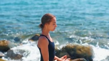 Siyah mayo giyen zayıf, bronz tenli kızın eğimli görüntüsü taşların üzerinde bacak bacak üstüne atarak oturuyor.