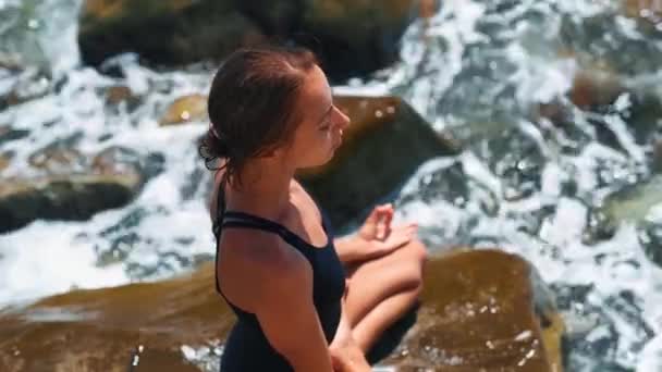 Cámara panorámica derecha vista superior de la mujer joven delgada en traje de baño sentado piernas cruzadas sobre piedras — Vídeo de stock