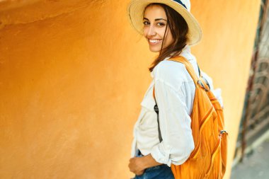 Hasır şapkalı çekici turist kadın, kameraya gülümseyen beyaz gömlek, turist şehrinde parlak turuncu duvara karşı duran..