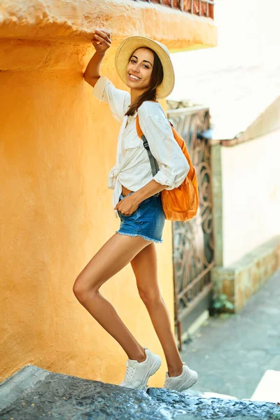 Ritratto a figura intera donna turista sorridente in cappello di paglia, camicia bianca posa contro parete arancione brillante in città turisti . — Foto Stock