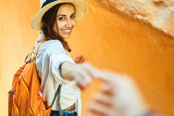 Красивая улыбающаяся туристка в соломенной шляпе, белой рубашке и оранжевом рюкзаке держа за руку своего парня, следуйте за мной . — стоковое фото