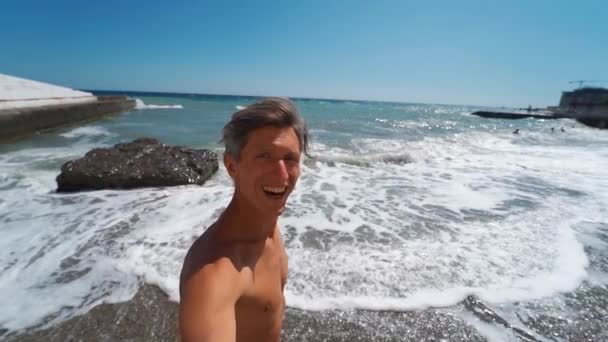 Heureux rire bronzé sportif jeune homme prendre vidéo selfie tout en marchant le long de la plage avec la mer ondulante — Video