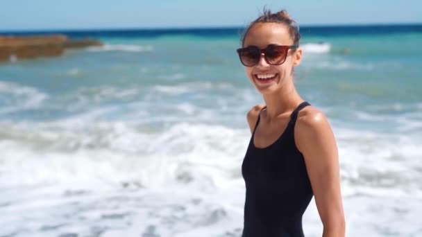 Alegre riendo sexy chica delgada en traje de baño negro y gafas de sol de pie en hermosa playa de mar soleado con olas . — Vídeo de stock