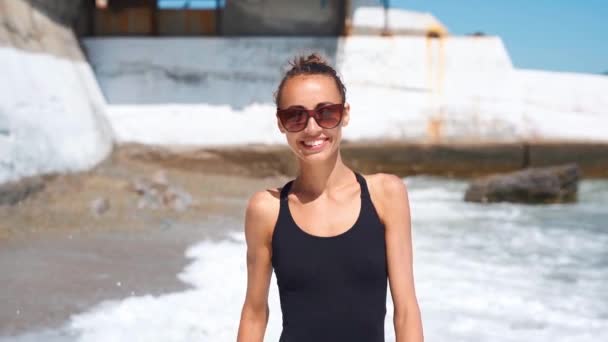 Медленное движение стройная сексуальная фигура, улыбающаяся молодая женщина в черных купальниках, идущая вдоль морского пляжа с волнами. — стоковое видео