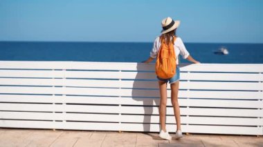 Yavaş çekim güzel kadın turist güneş şapkası, gömlek ve şortla deniz manzarasının tadını çıkarıyor.