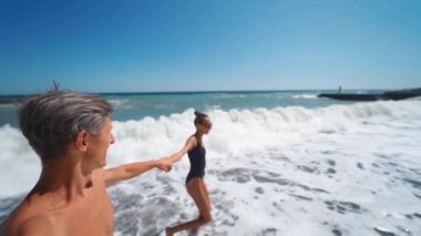 Geniş açı ağır çekim mutlu çift el ele tutuşup dalgalı deniz sahillerinde neşeyle yürüyor.