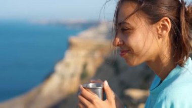 Uçurumun kenarında oturan, kahve ya da çay içen genç bir kadın. Burun ve deniz manzarasının tadını çıkarıyor..