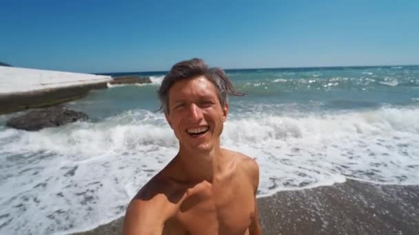 Улыбающийся спортсмен делает селфи, стоя на пляже с волнистым морем — стоковое видео