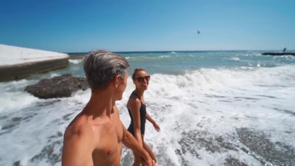 Glückliches Paar Händchen haltend spaziert freudig am winkenden Meeresstrand — Stockvideo