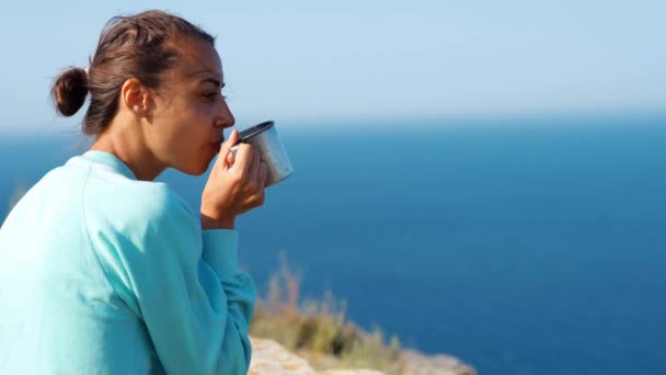 Портрет на открытом воздухе беззаботная улыбающаяся женщина сидит на краю скалы с прекрасным видом на море, держа кружку кофе или чай в руках — стоковое видео