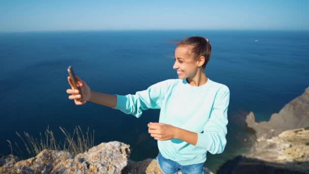 Młoda kobieta podróżnik co selfie na krawędzi klifu z pięknym widokiem na morze, śmiech i uśmiech do kamery, co zabawne twarz. — Wideo stockowe