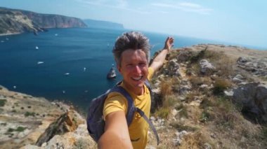 Genç erkek turist, uçurumun kenarında özçekim yapıyor ve gülümsüyor. Güzel deniz manzarası ve harika doğanın tadını çıkarıyor..