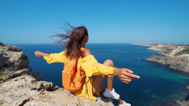 Nahaufnahme Porträt einer gebräunten Reisenden, die mit erhobenen Händen, leuchtend gelbem Kleid und im Wind wehenden Haaren auf einem Felsstrand sitzt. — Stockvideo