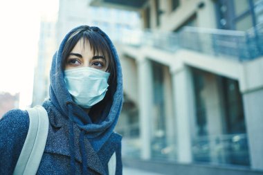 Şehirdeki koronavirüsü korumak için maske takan kapüşonlu bir kadın. Arka planda modern bir bina var..