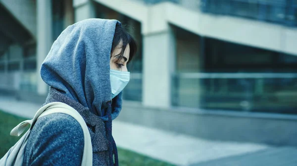 Side view woman in hood with face mask for protection of coronavirus with modern building on background. — Stock Photo, Image