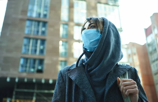 背景に建物と街の通りに立って、予防コロナウイルスのための保護顔マスクを身に着けてフードの女性. — ストック写真