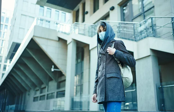 woman in hood with backpack standing against modern building in face mask for protection of coronavirus, looking to camera.