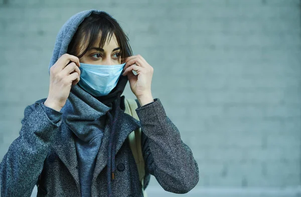 灰色の壁に立っているコロナウイルスの保護のために顔のマスクを身に着けているフードの女性. — ストック写真