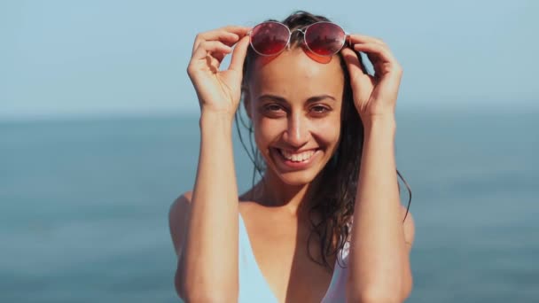 Buiten portret van Mooie stijlvolle sexy sportieve vrouw in zwart badpak en witte pet wandelen langs baai aan zee, tegen een blauwe zee op zonnige dag. — Stockvideo