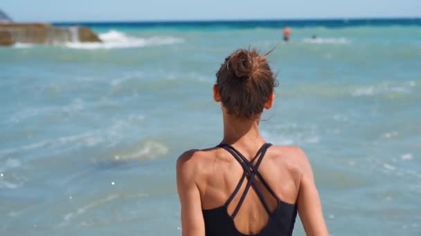 Visão traseira câmera lenta esbelta jovem em roupa de banho preta em pé contra o oceano azul — Vídeo de Stock