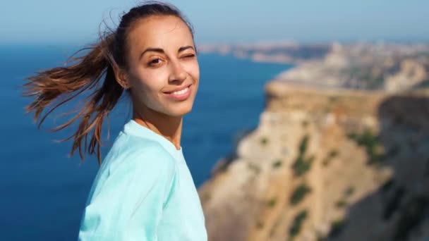 Retrato de la joven feliz de pie en el borde del acantilado contra el increíble paisaje marino y el cielo, pelo que sopla en el viento — Vídeo de stock