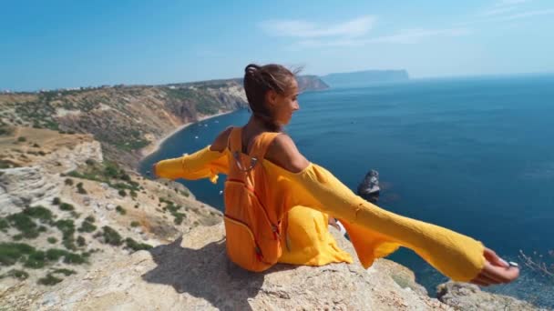 Mulher alegre viajante vestido amarelo sentado penhasco borda levantando as mãos desfrutando da paisagem marinha — Vídeo de Stock